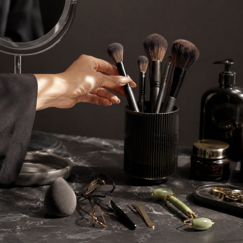 Makeup brushes in a black holder on a dark surface with a hand reaching for one of the brushes.